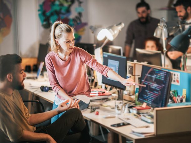 Woman sitting on desk pointing at computer screen to a developer and collaborating.